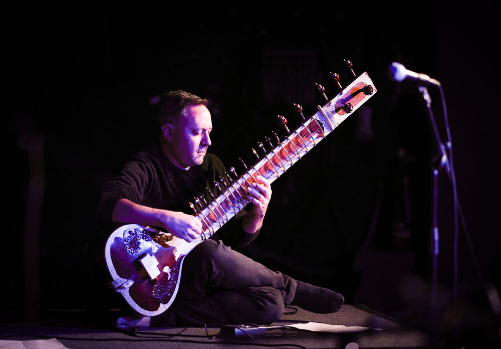A man sitting cross legged on a stage playing the sitar