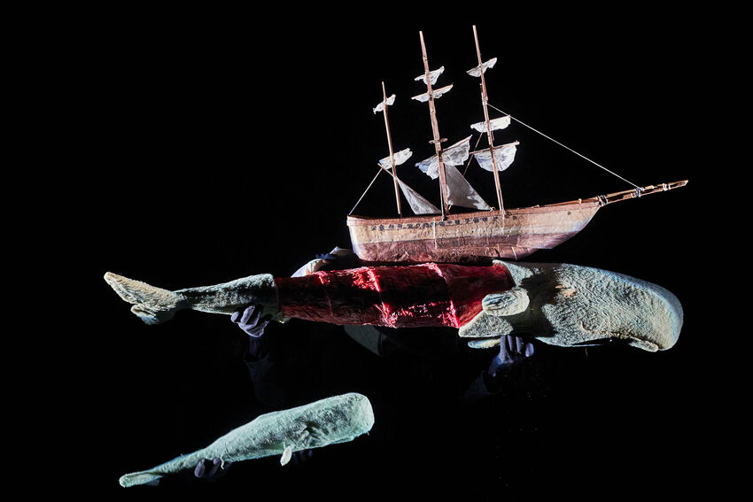 A image of two very large whale puppets on a black stage. One appears to be swimming underneath a sailboat