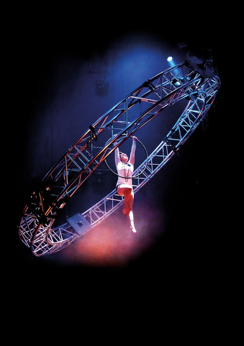 An acrobat hangs from a hoola hoop within another larger ring