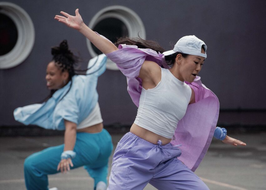 In the foreground a person wearing light purple and a white backwards cap dances, arms outstretched. Behind a person also dances, arms outstretched. They are wearing light blue.