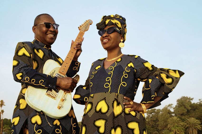 Amadou (left) with a guitar wearing sunglasses and a suit with yellow heart patterns and Mariam (right) in sunglasses, a yellow heart pattern dress and headscarf, a clear blue sky behind them