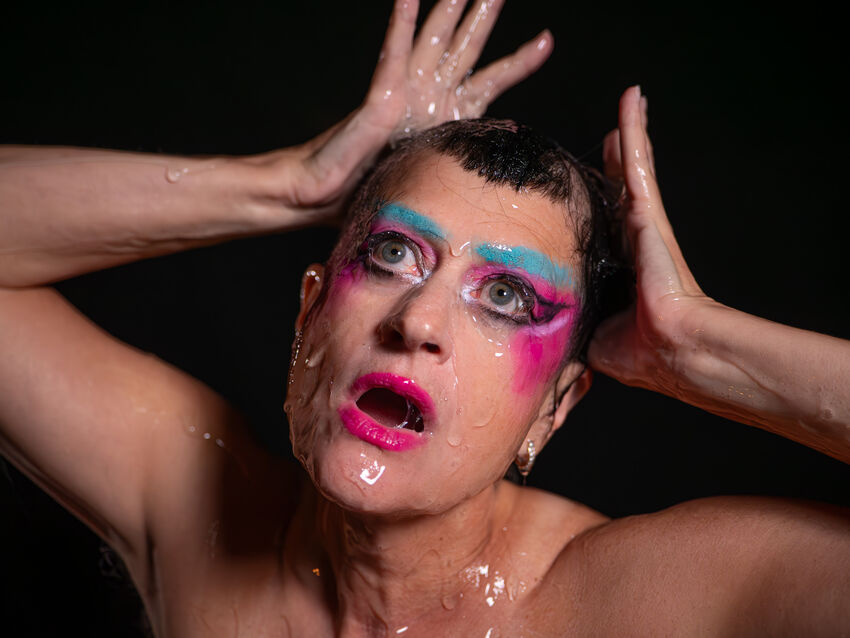 Peaches holding her hands to her hand and wearing pink blush, eyeshadow and lipstick. She has bright blue eyebrows and is covering in a clear slippery liquid