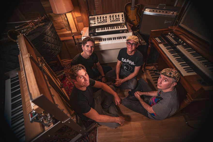 Four men sit cross-legged on the floor surrounded by pianos of various sizes. They're looking up towards the camera