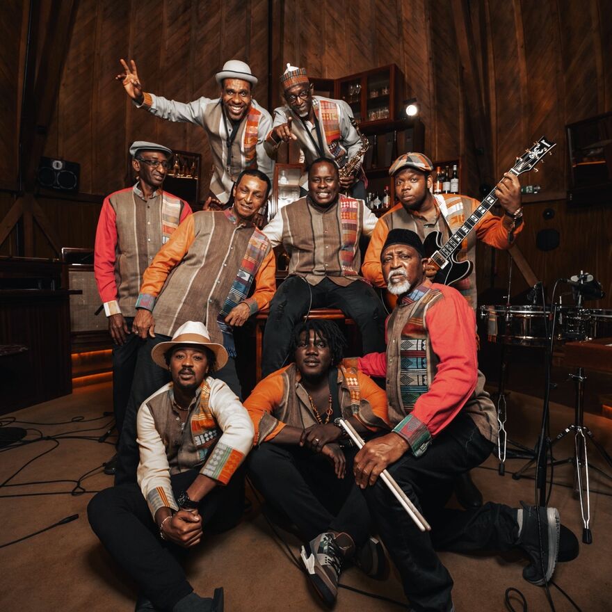 A group of black musicians all pose in an old building with their instruments. They are all wearing bright colours and African inspired prints.