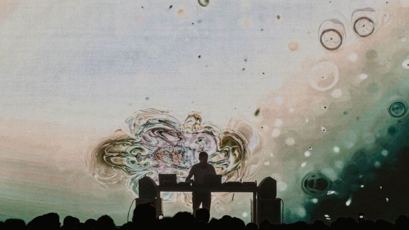 Max Coper at a DJ deck, a screen projecting organic blob shapes of white, cream and teal 