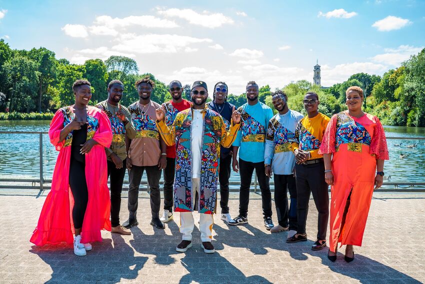 Seckou Keita and the Homeland Band pose next to a river and trees on a sunny day, all wearing bright colours and patterns