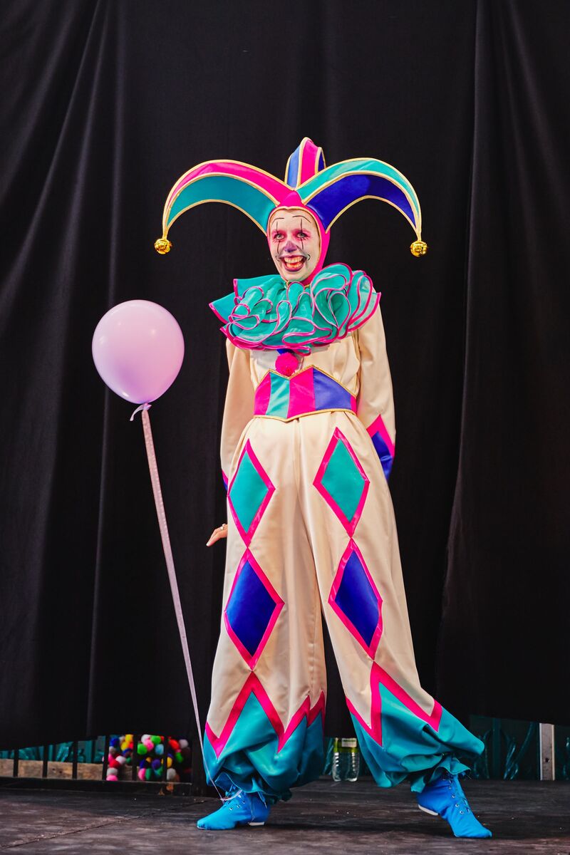A person dressed in a pink, turquoise and blue jester-style clown costume, with a diamond pattern on the trousers and a cap with bells