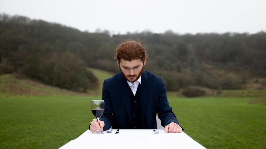 A young man with facial hair sits at a table clutching a wine glass which is half full of red wine. He looks down at the table. He is in a green field with hills in the background