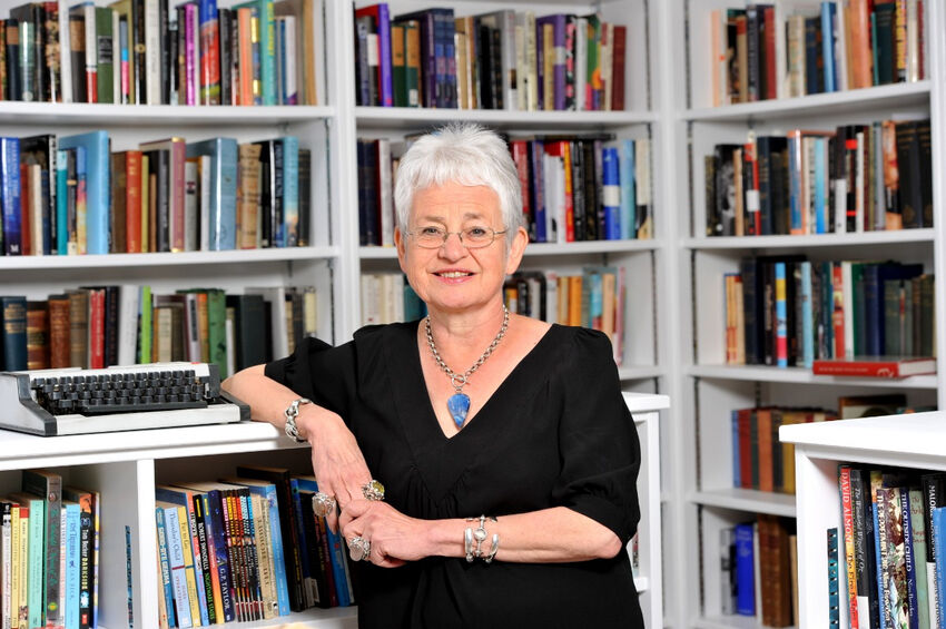 Jacqueline Wilson sitting in front of a wall-to-wall bookshelf. Jacqueline Wilson has white hair, glasses, and is wearing a black top and chunky blue and silver necklace