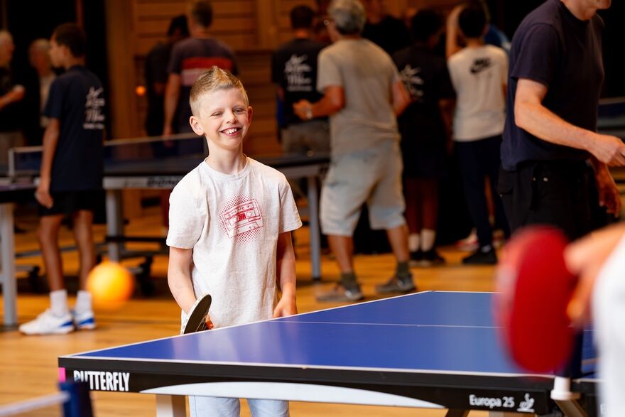 A young boy stands at a table tennis table holding a table tennis bat. He is wearing a white t-shirt and is smiling and enjoying himself. In the background are lots of people of all ages playing table tennis
