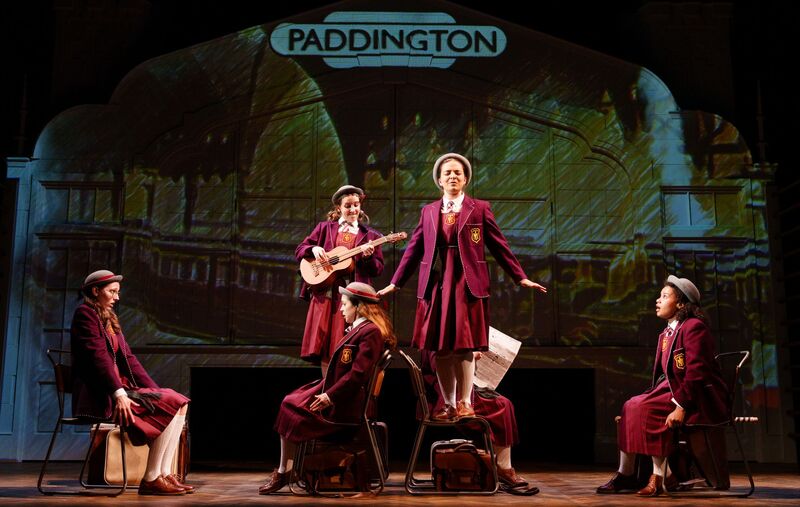 Five people in school uniforms. Three sitting on chairs, one standing on a chair and another playing the guitar on stage