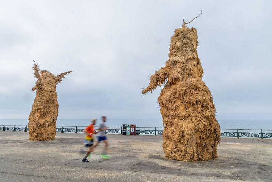 Two large sculptures made of agricultural materials on Hove Promenade, with two blurred runners in the foreground