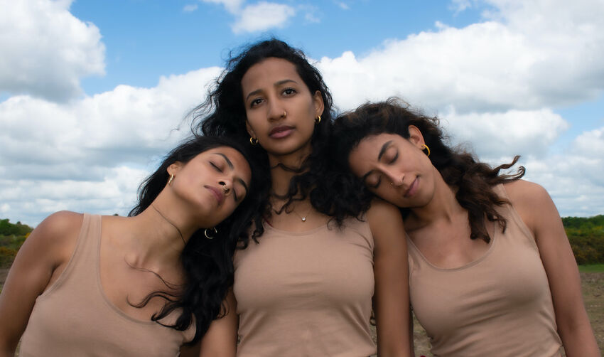 Three women post with their heads together. They all have long dark hair and wear flesh coloured vest tops. They are outside and in the background there is a clear blue sky and rolling hills