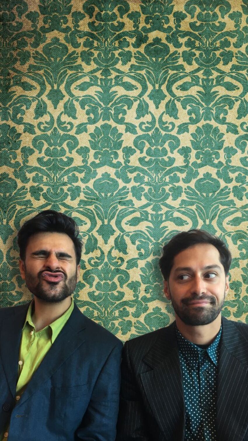 Two men stand in front of a yellow and green patterned background. They are both facing towards the camera, one is pouting, and the other is looking towards him