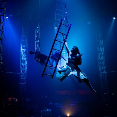 A woman holding on to a ladder, that is suspended in air. Behind her are two other people holding on to suspended ladders. They are all lit in a blue light.