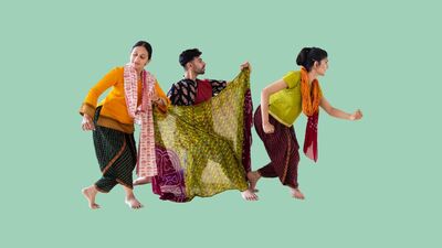 Three dancers dressed in traditional Indian clothing pose and gesture towards the right of the frame. They are against a plain green background