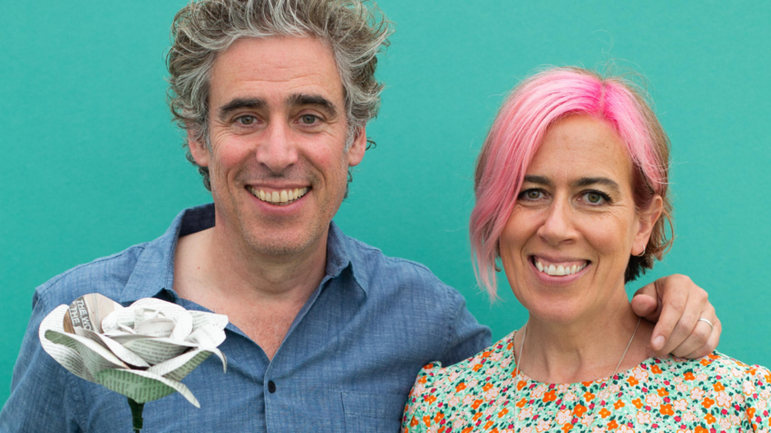 Stephen and Anita Mangan stand together smiling. Stephen has his arm around Anita and he is holding a large white rose.