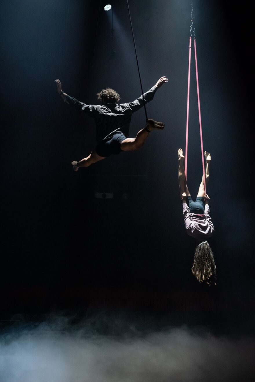 One performer flies through the air whilst another hangs from two red ropes, upside down