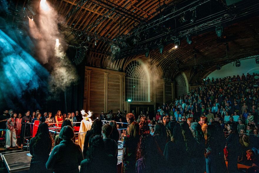 A performer stands in the middle of a boxing ring with an audience surrounding them