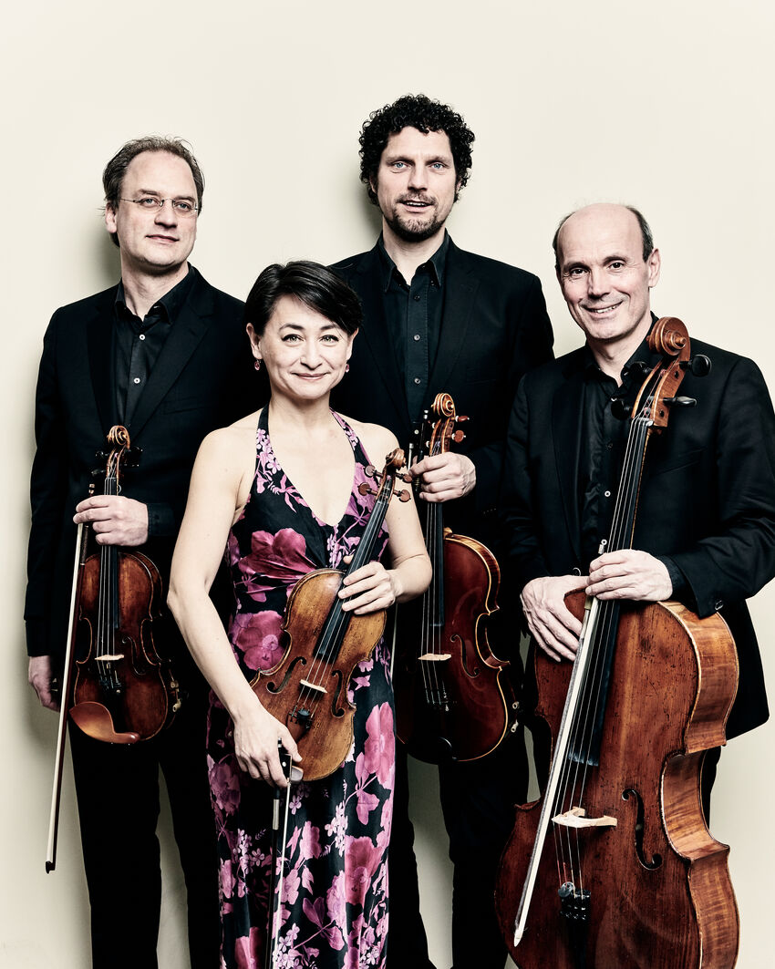 Ruysdael Quartet standing two front, two behind. Back left, a man with short brown hair wearing a black suit holding a violin. Back right, a man with black curly hair wearing black suit, holding a viola. Front left, a woman with black hair and a side fringe wearing black halter dress with pink flowers holding a violin. Front right, a man with dark hair around the sides wearing a black suit holding a cello