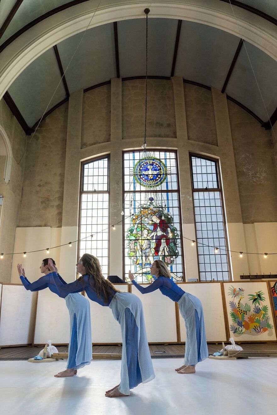 Three female dancers in fully blue outfits stand, slghtly leant forward with their arms raised. Behind them is a large stained glass window