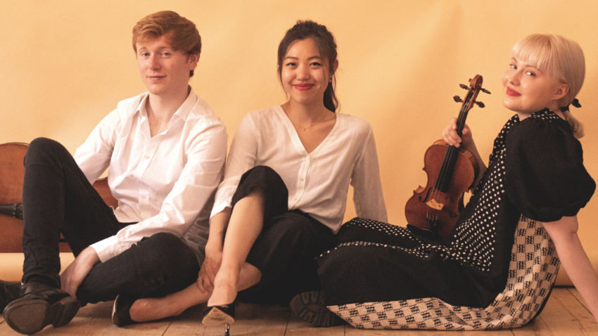 Members of Paddington Trio recline with instruments in front of a yellow wall