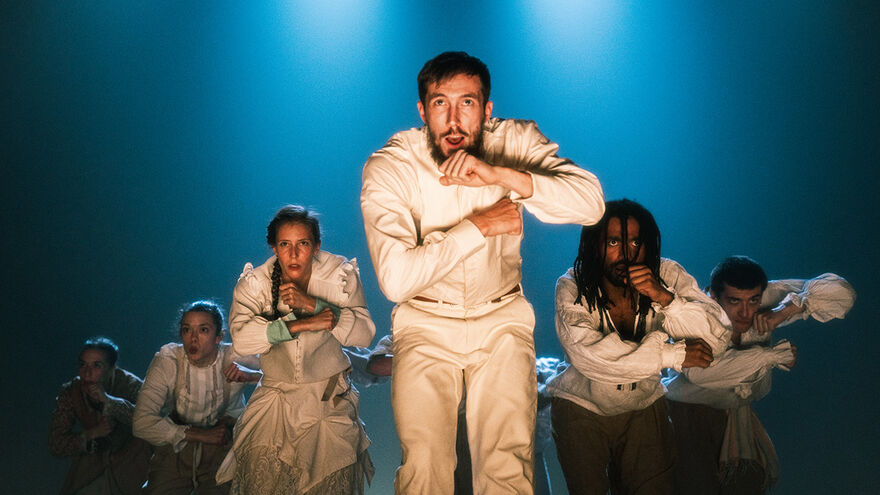 A male dancer at the front of a V-formation of six other dancers. All wearing white and crouching slightly, with a blue background