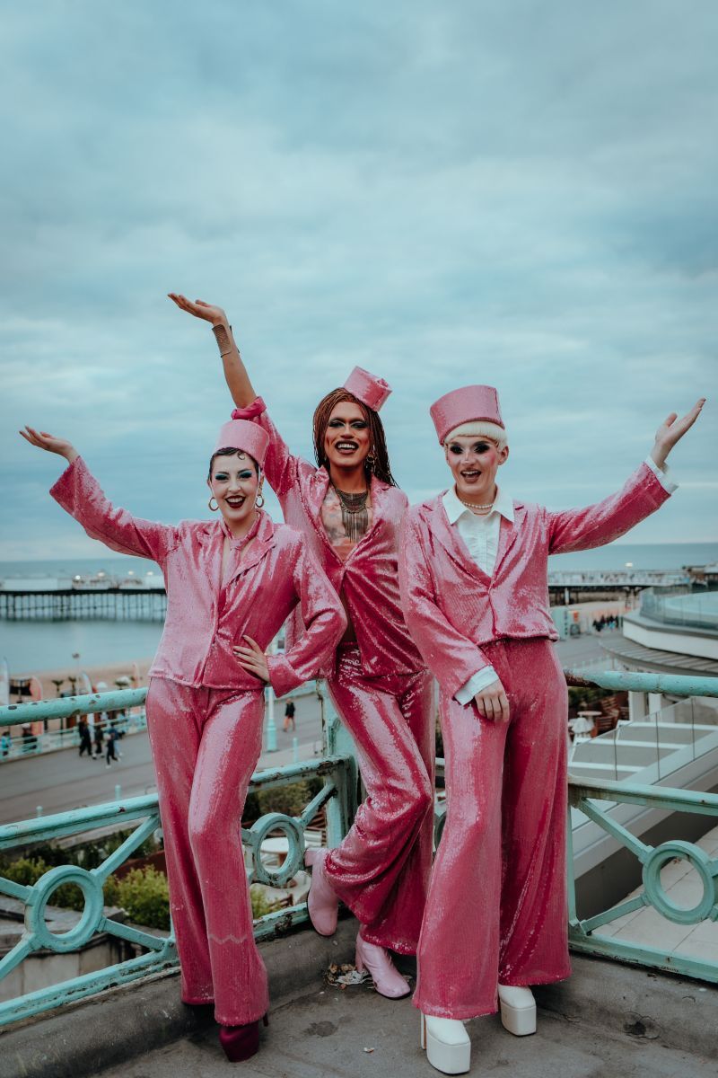 Billie Gold, Alex Fincher and Alfie Ordinary pose with their hand in the air, wearing pink sequin suits and hat, in front of the green seafront railings with the beach behind