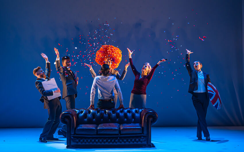 Six performers on a blue stage behind a leather couch throwing confetti in the air