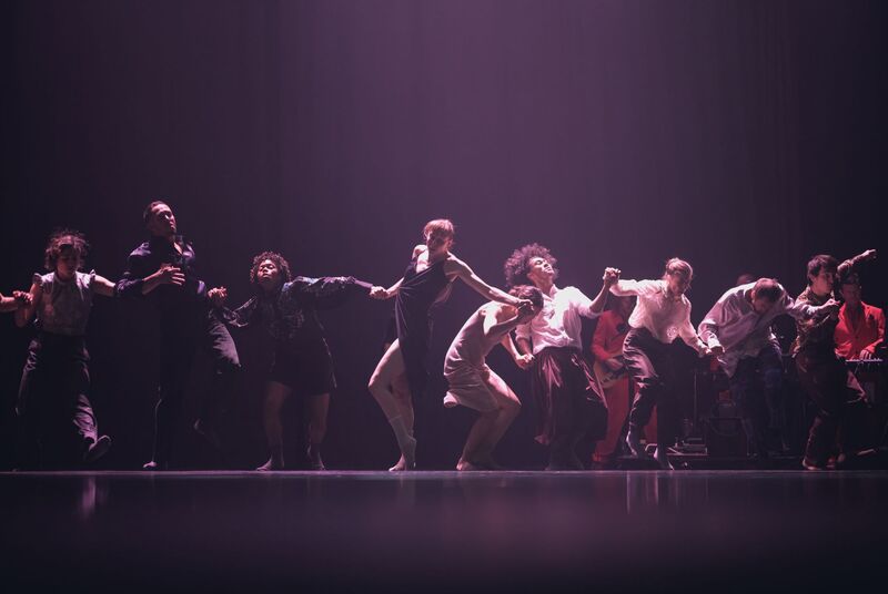 9 dancers stand in a line stretching across a dark stage, holding each other’s hands creating a chain of bodies. Each dancer is positioned differently, with some holding there arms high and thrusting their bodies outwards, and others hunched over. They all seem to be in their own world, not looking at each other. The dancers are wearing an assortment of different colourful shirts, trousers and dresses. Behind them - on the right of the image - 3 musicians stand, playing various instruments. They are dressed in red suit trousers and red blazers.
