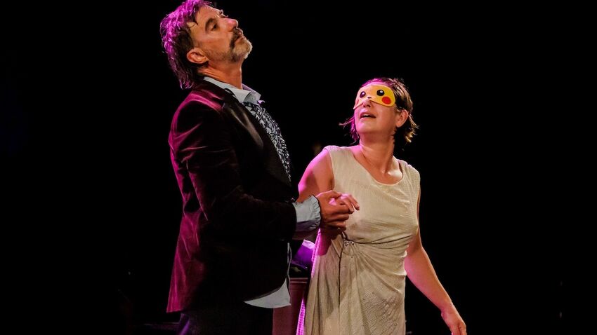 A man and a woman hold hands on stage, the woman is wearing a blindfold