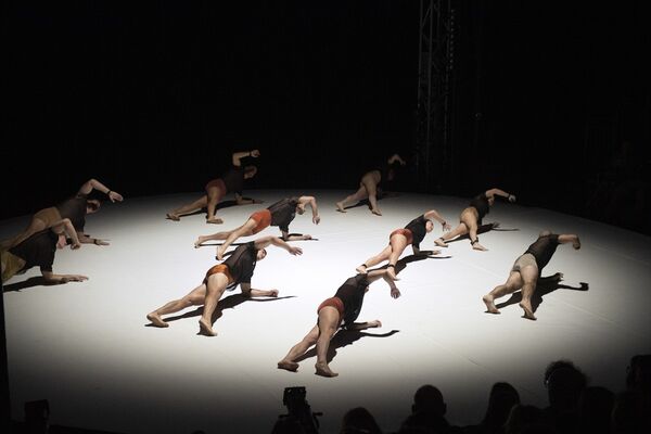 In a large white spotlight, a group of acrobats lay on the floor, all with their right arms and half of their bodies lifted off the floor