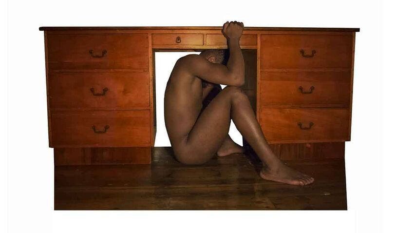 A black man crouches naked under a desk
