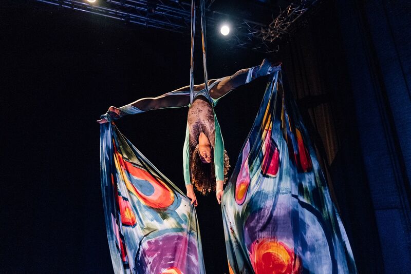 A young girl of colour hangs from some ropes, her legs spread far apart, there are big pieces of colourful material hanging from her feet and held in her hands