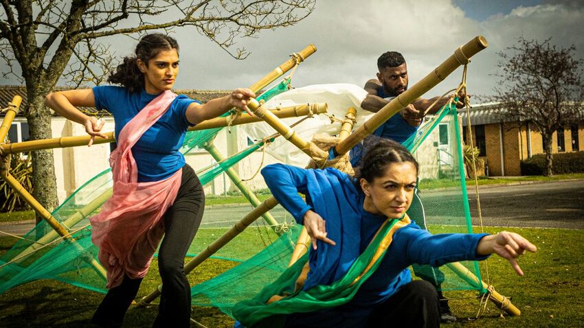 Three dancers dressed in blue gesture towards the left side of the image. Their hands perform mudras and they are surrounded by bamboo poles and green netting 
