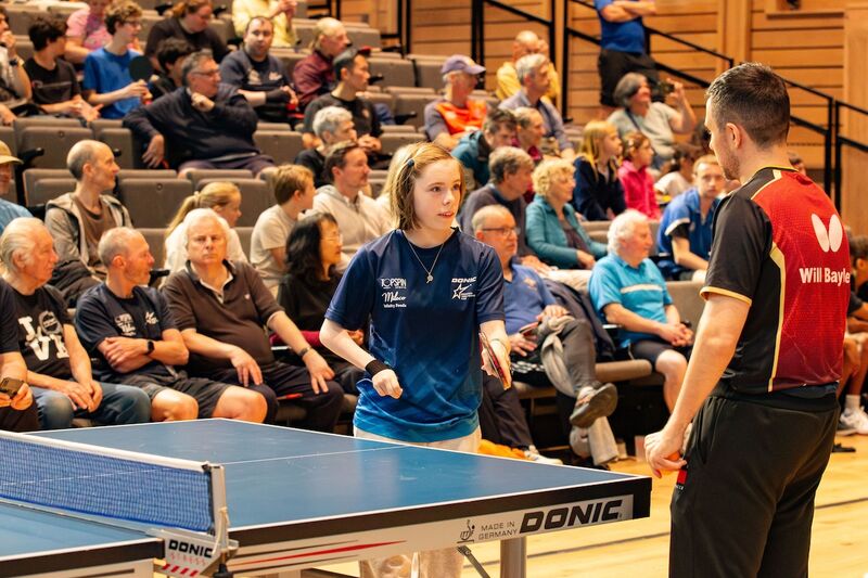 A female presenting table tennis player is getting coached by Paralympic table tennis star Will Bayley. In the background, people watch on from terraced seating 
