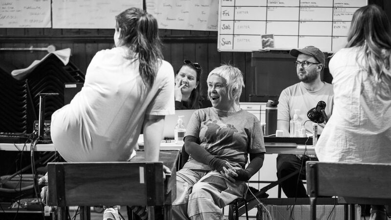 Two girls with their backs to the camera sit on top of school desks while a woman at the front smiles towards them