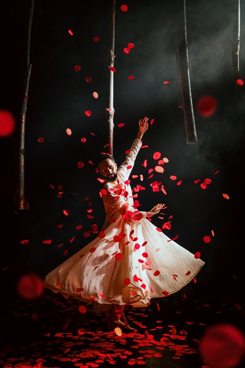 A man in a white dress, with arms held up to his left, he's dancing as red petals fall around him
