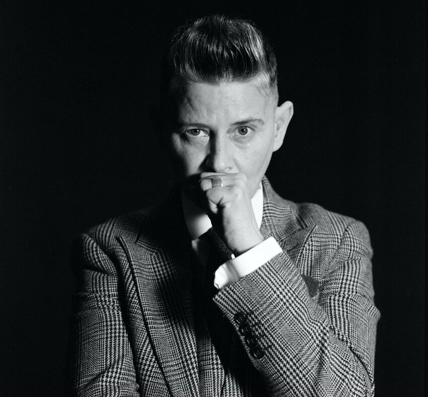 A portrait of Joelle Taylor in greyscale. She looks directly at the camera with one of her hands in a fist resting on her mouth