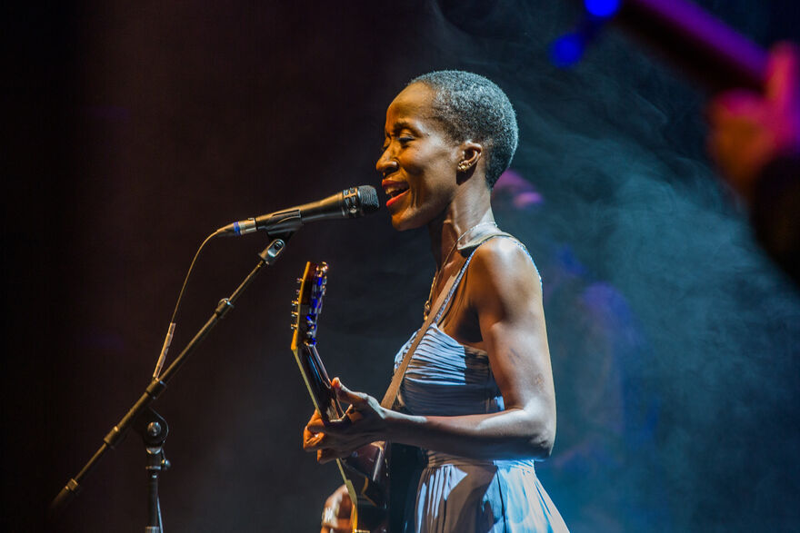 Rokia Traore holding a guitar and singing and smiling, with a microphone in front of her at event Ne So at Brighton Festival
