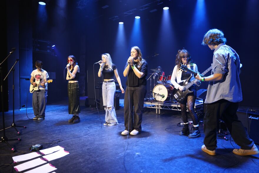 Seven young musicians on a stage, some singing with microphones, some playing guitar and another on drums. They are lit with blue backlights.