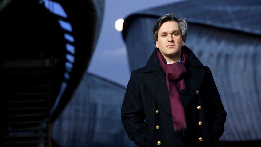 A white man with grey hair stands in front of some grey, curved buildings. He wears a black coat and purple scarf