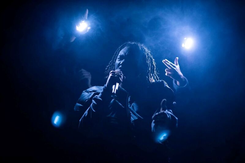 A man with shoulder length hair is lit from behind by bluie lights. He is shouting into a microphone making gun signs with his hands.