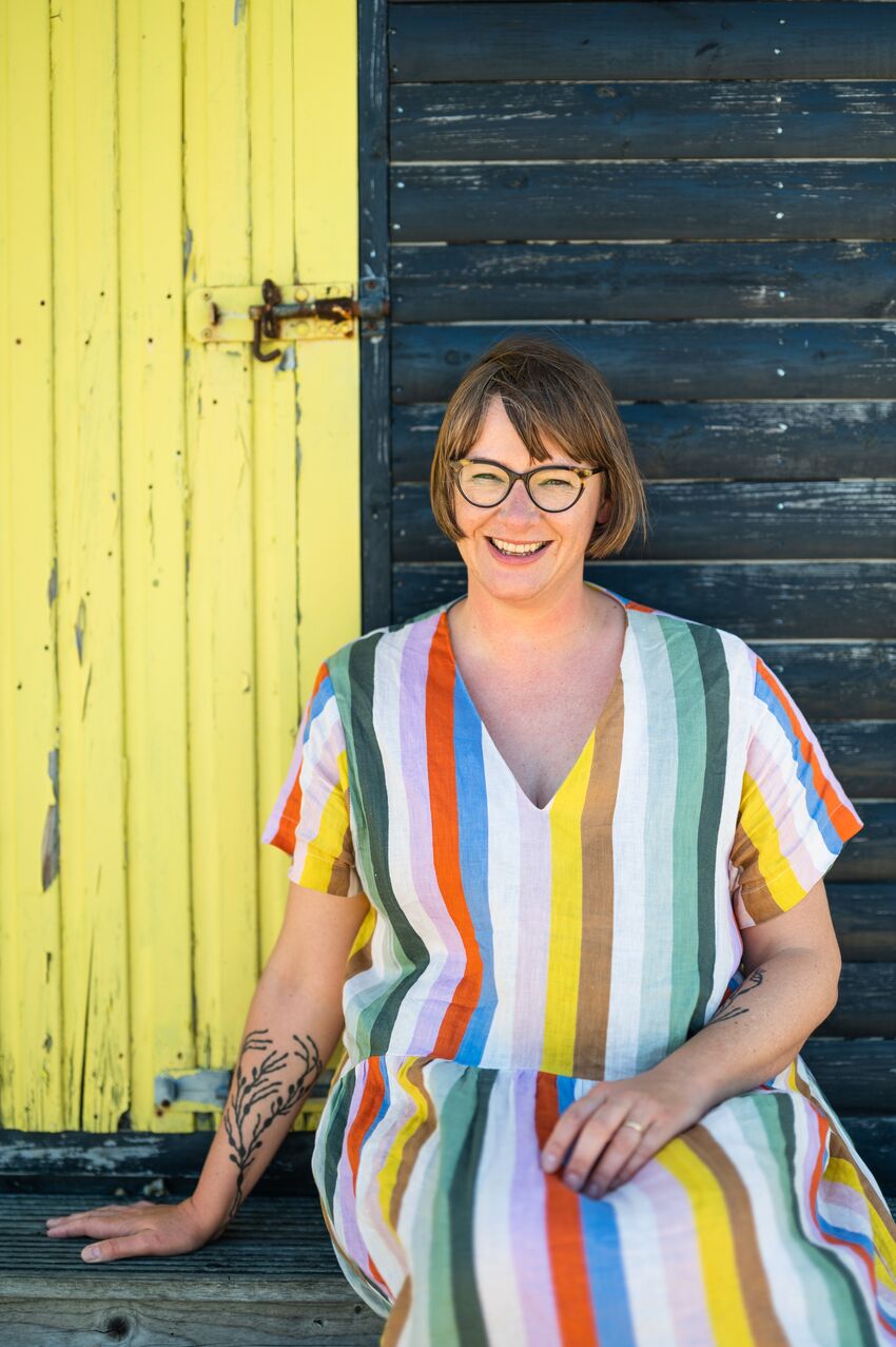 Katherine May wearing a colourful striped dress smiling next to a rustic wooden door