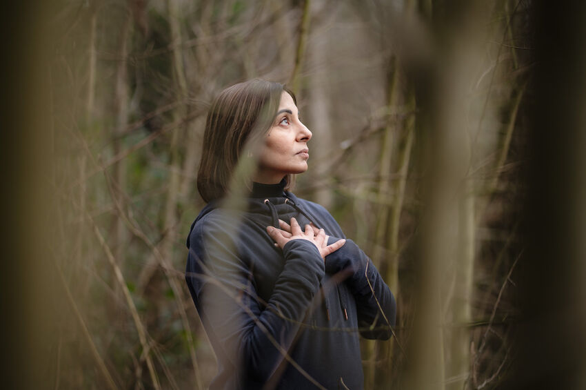 Parmjit Sagoo stands in a forrest. She wears a dark jumper and has her hands on her chest. She is deep in though