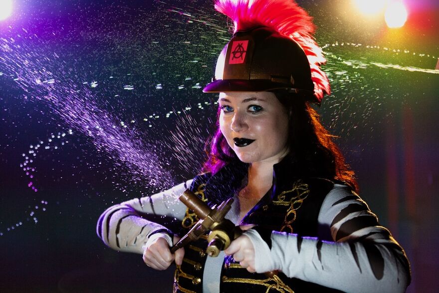 A woman stands looking at the camera. She wears a hard hat, and holds a hose that is spraying water