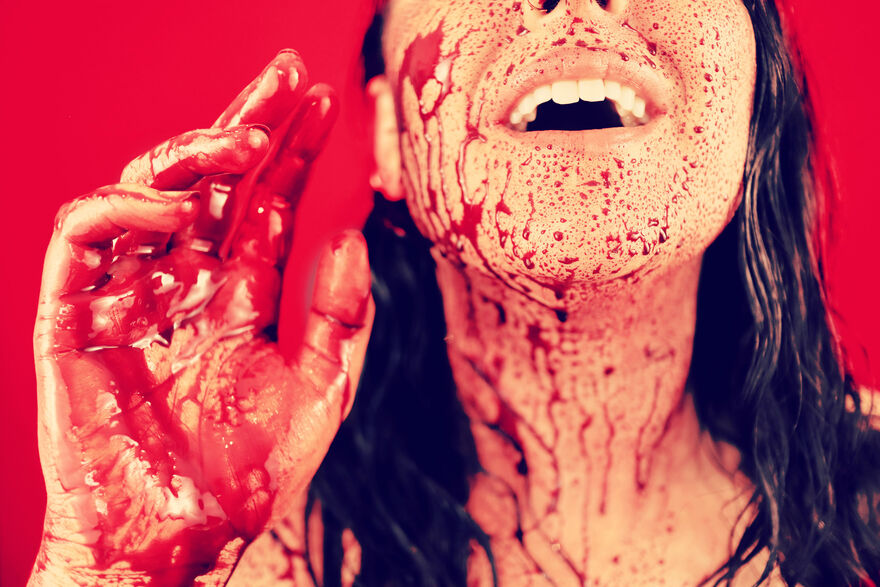 The bottom half of a persons face, and their neck is covered in blood, as is their hand that they're holding up