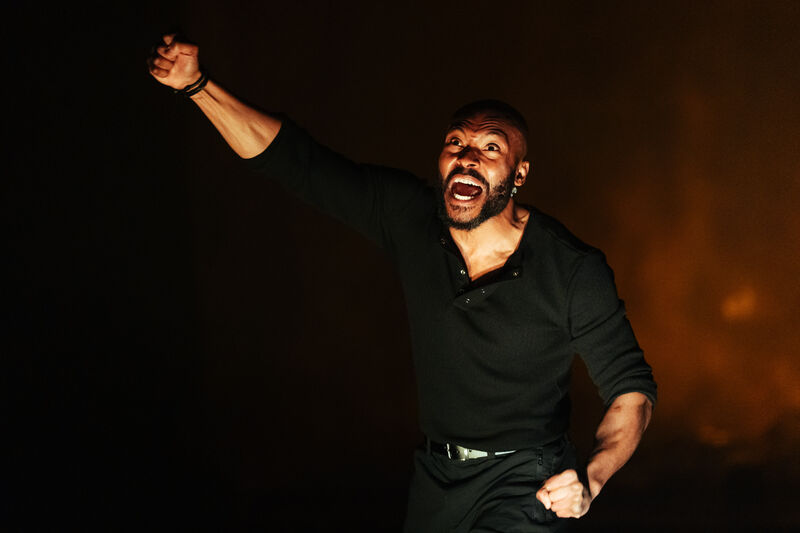 A man with his fist in the air screaming. He is lit with an orange light and surrounded by haze. He looks angry