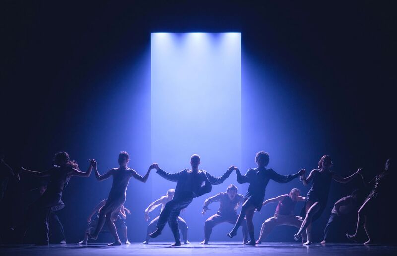 13.	A Large group of 12 or 13 dancers stand on a stage in 2 lines going from left to right - one line of dancers in the foreground and another line of dancers directly behind them. The foremost line of dancers are all holding hands, and each is posting on their right leg and swaying their body to the left while kicking their left leg out to the right. The line of dancers behind them are crouched down low with their legs splayed wide, and their arms held out to each side, curved downwards at the elbow. The first line of dancers look innocent, dancing together happily, whilst the line behind them stare at them animalistically. They are dressed in an assortment of colourful shirts, trousers and dresses.