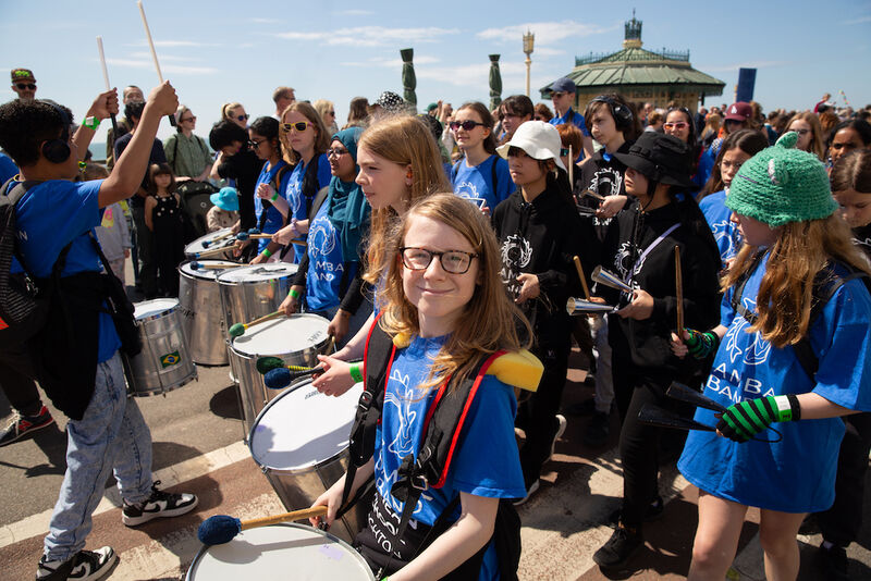 A group of children are parading along the seafront carrying drums as a samba band
